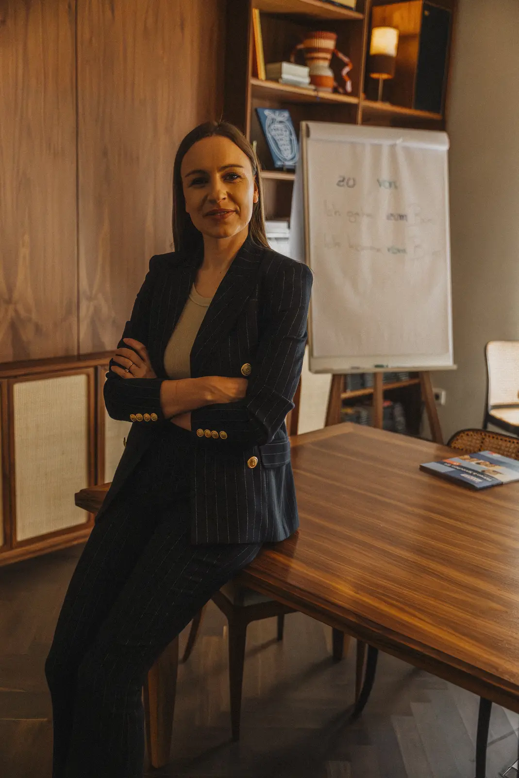 Lena in a navy suit, arms crossed, leaning on a wooden table